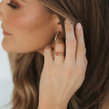 A close-up lifestyle shot of a woman wearing a delicate gold beaded bracelet on her wrist, a gold hoop earring, and a gold ring with small red gemstones, set against her brown hair and tan top.
