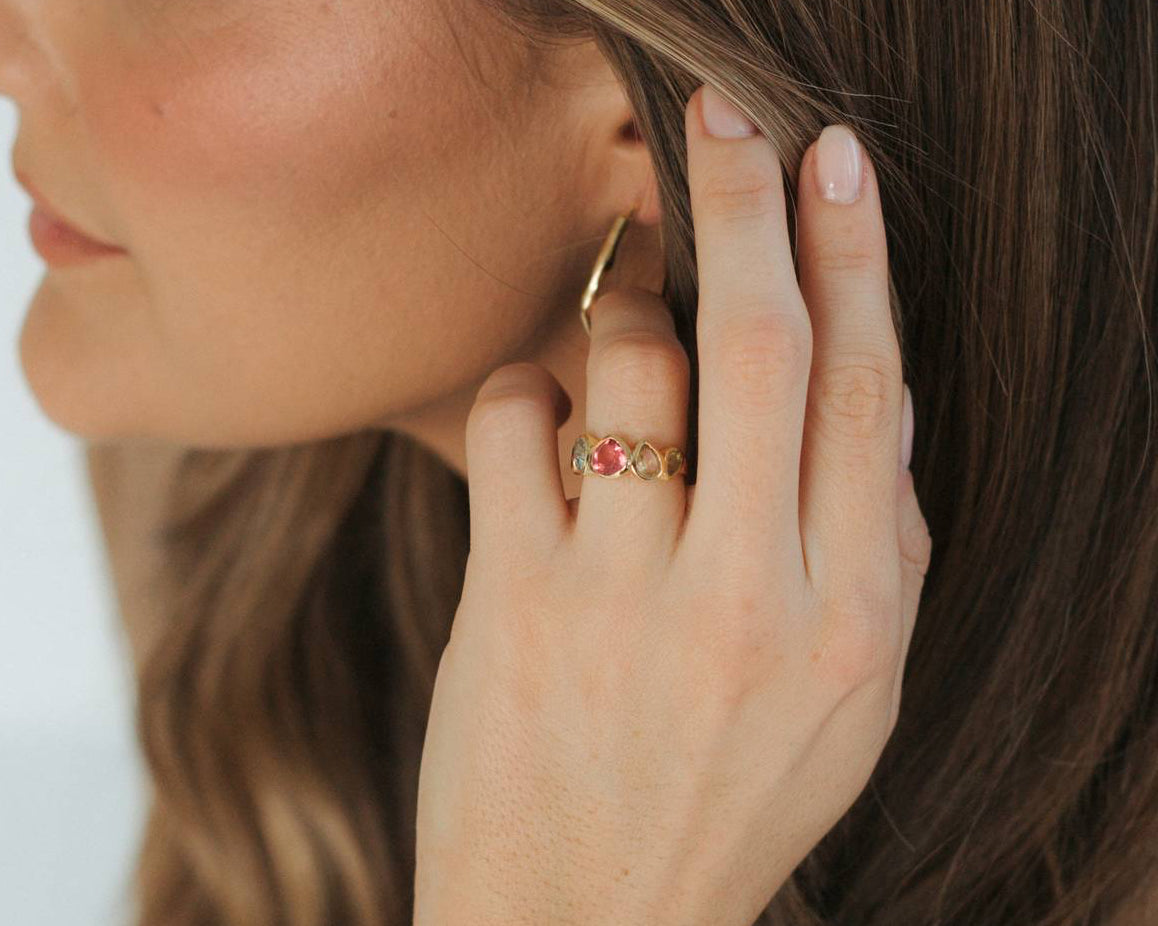 A close-up lifestyle shot of a woman wearing a delicate gold beaded bracelet on her wrist, a gold hoop earring, and a gold ring with small red gemstones, set against her brown hair and tan top.