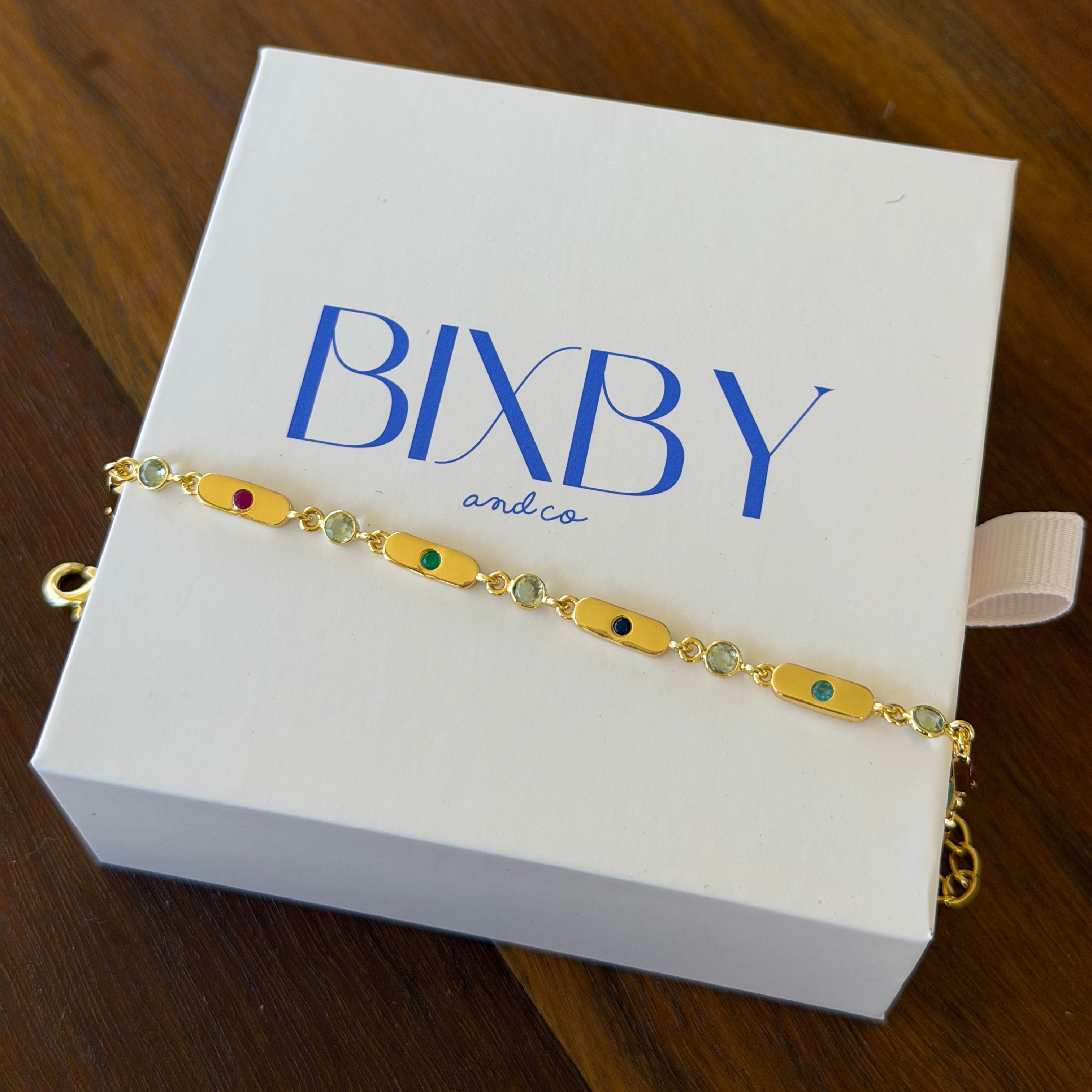 A gold-toned beaded bracelet with colorful oval beads lies across a white jewelry box printed with the brand name 'BIBY and co'. The box is placed on a dark wood surface.