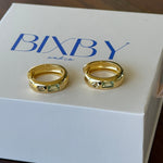 Two gold hoop rings displayed on a white BIXBY brand box, set against a dark wooden background.
