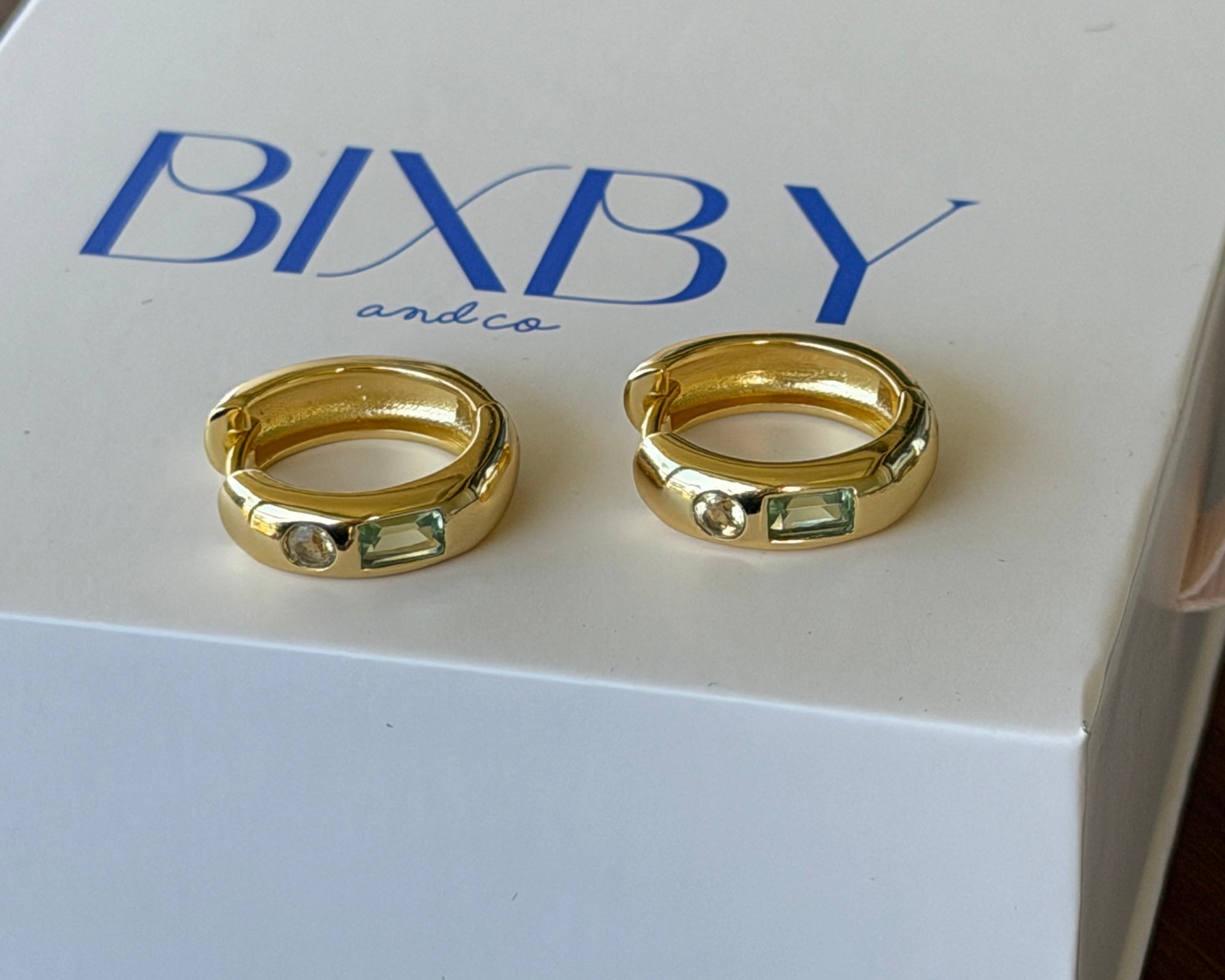 Two gold hoop rings displayed on a white BIXBY brand box, set against a dark wooden background.