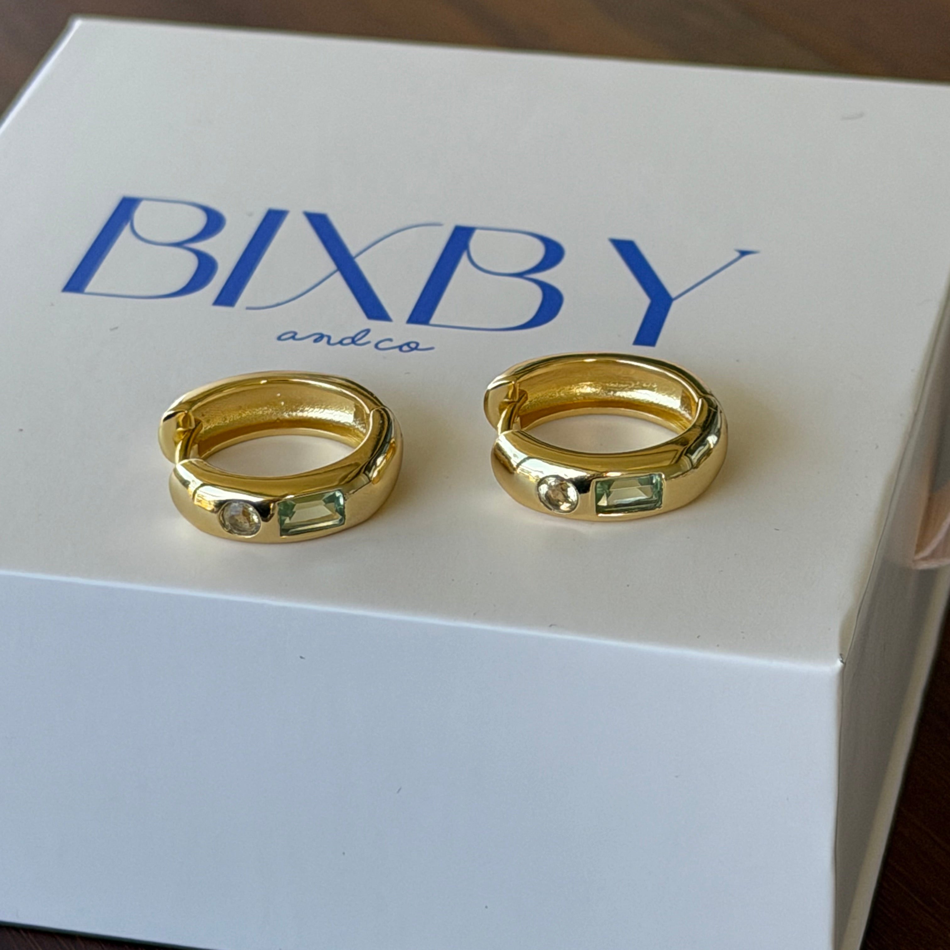 Two gold hoop rings displayed on a white BIXBY brand box, set against a dark wooden background.