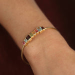 A gold-plated bangle bracelet with a row of small rectangular multicolor gemstones, shown worn on a person's wrist against a dark brown background.
