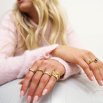 Close-up of a person's hands wearing multiple gold rings with a pink sweater and white background.