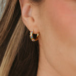 A close-up of a person's ear wearing a small gold crescent-shaped hoop earring adorned with colorful rhinestones.