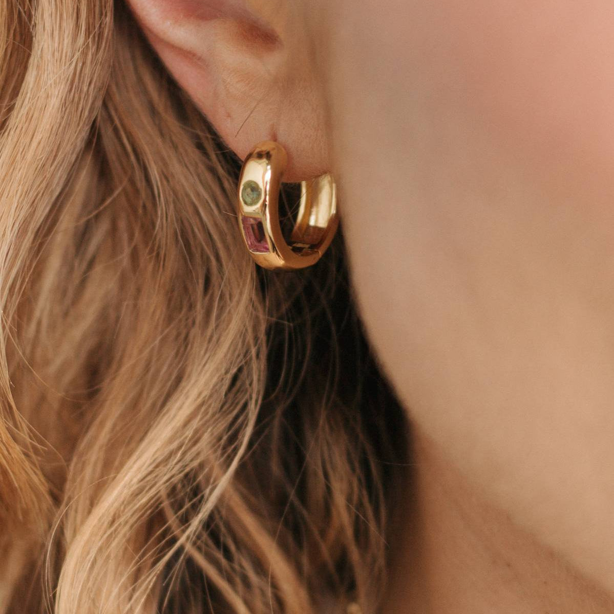 A close-up shot of a model's ear wearing a polished gold huggie hoop earring with a small green gemstone accent.