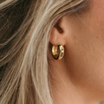 A close-up of a person's ear wearing a small gold hoop earring with clear stones, framed by blonde hair.