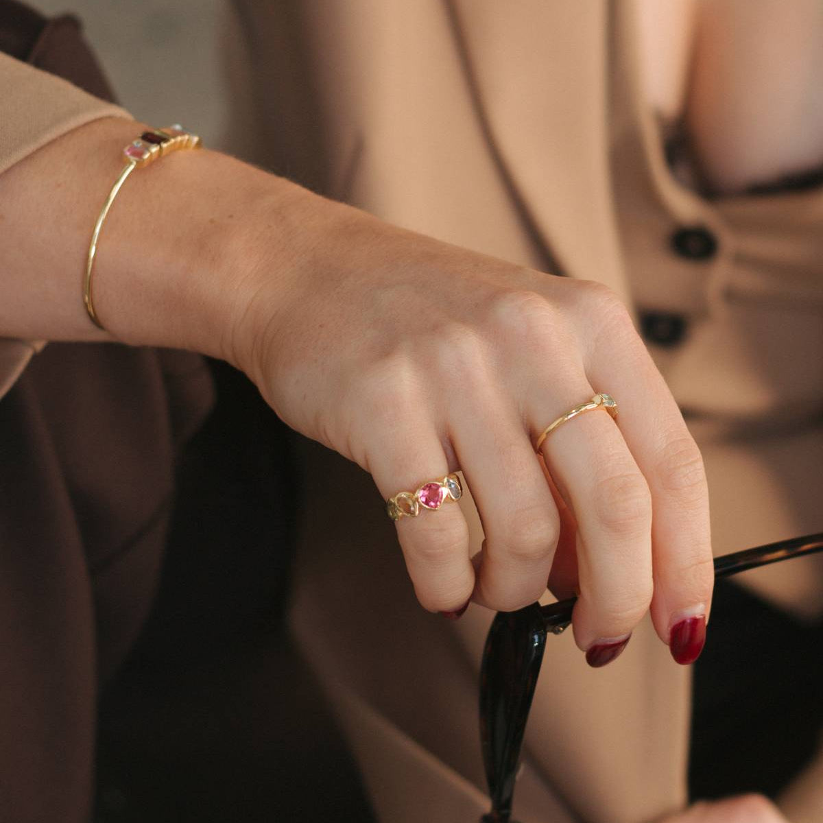 A close-up lifestyle shot of a person's hand wearing two gold rings, one with a small pink gemstone, along with a gold bangle bracelet and a beige blazer.