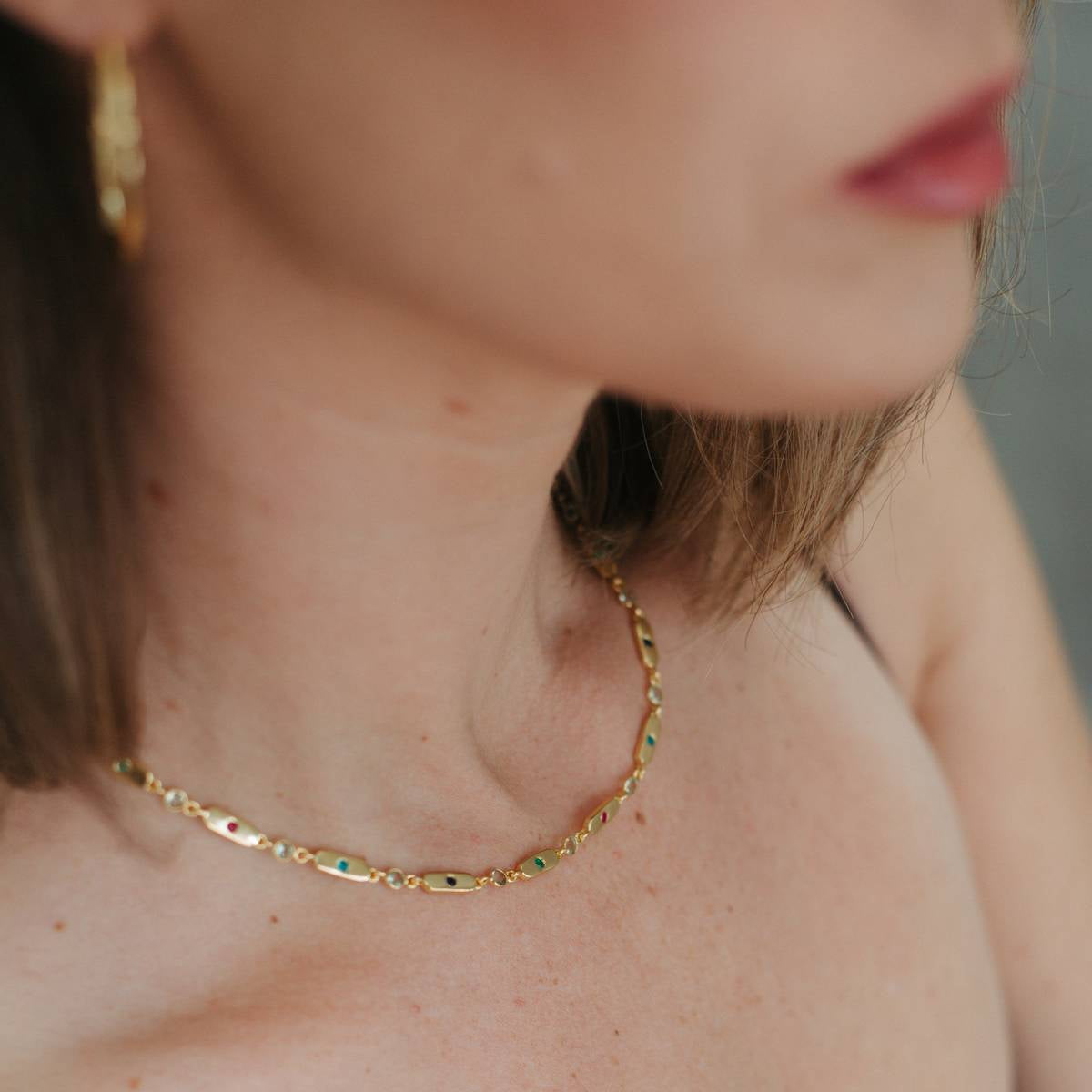 A close-up lifestyle shot of a woman wearing a delicate gold chain necklace with small green and blue stones, paired with a black spaghetti strap top.