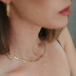 A close-up lifestyle shot of a woman wearing a delicate gold chain necklace with small green and blue stones, paired with a black spaghetti strap top.