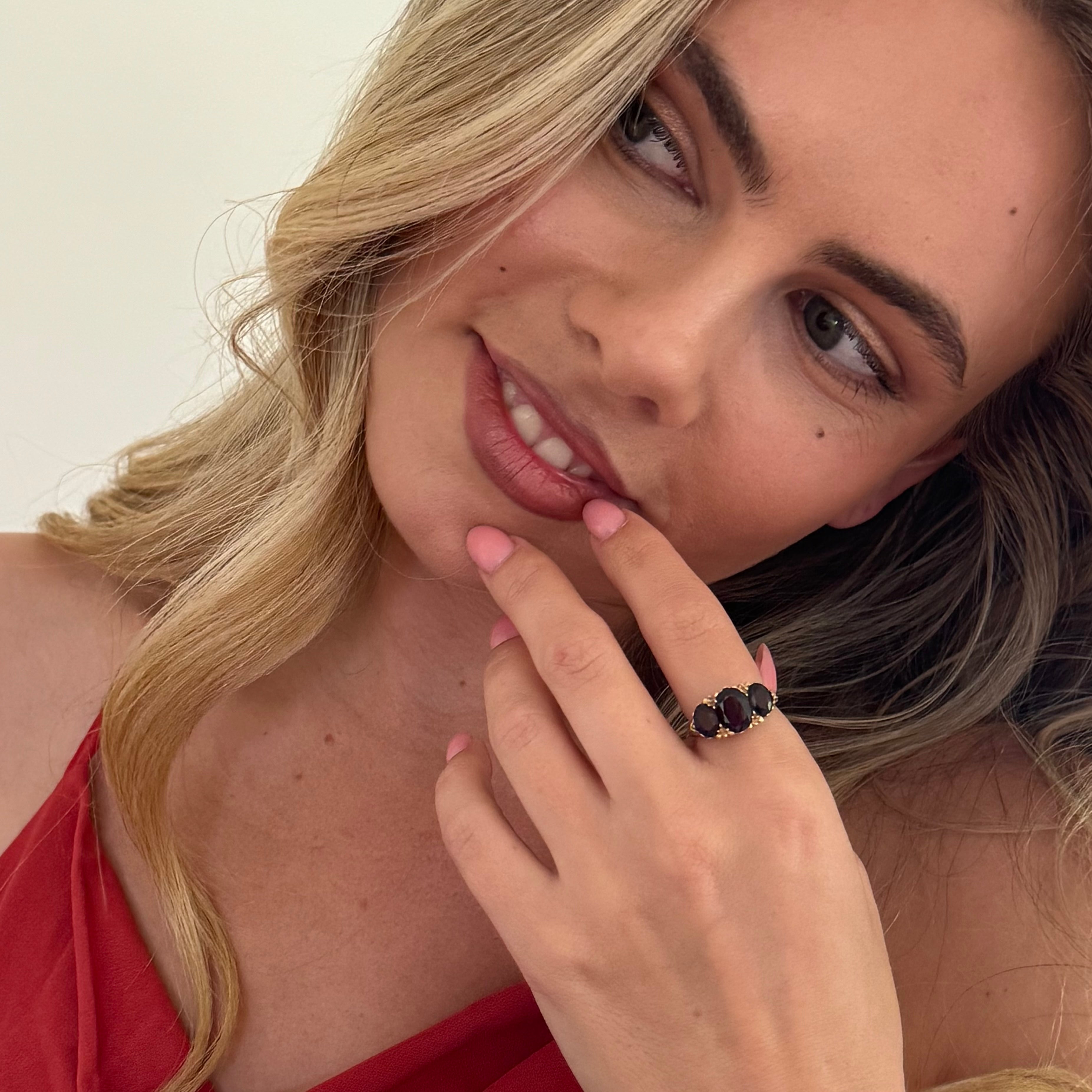 Woman with a red top and a ring on her finger, posing with one hand near her face.