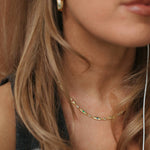 Close-up of a woman wearing a delicate gold chain necklace with small green stone accents, along with a gold hoop earring and white earbud wire.