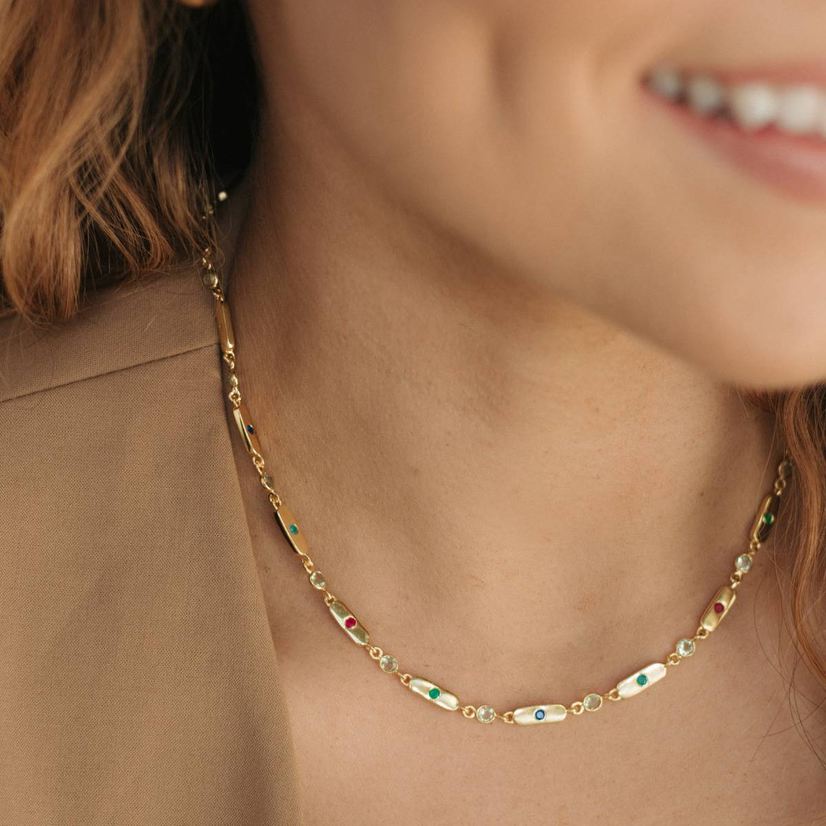 A close-up lifestyle shot of a woman wearing a delicate gold chain necklace with small oval charms featuring green, red, and white inlay details, paired with a gold hoop earring.