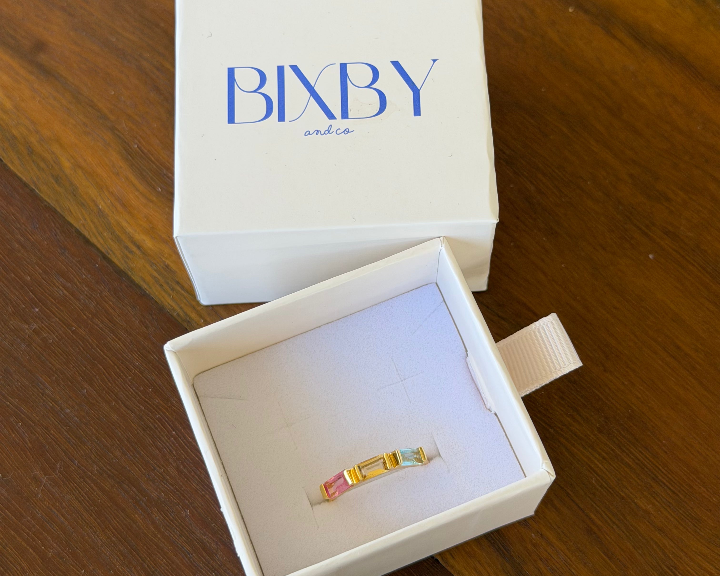 A gold ring with colorful enamel details displayed inside an open white jewelry box, with the white lid featuring the blue 'BIXBY' brand logo placed behind it on a wooden table.