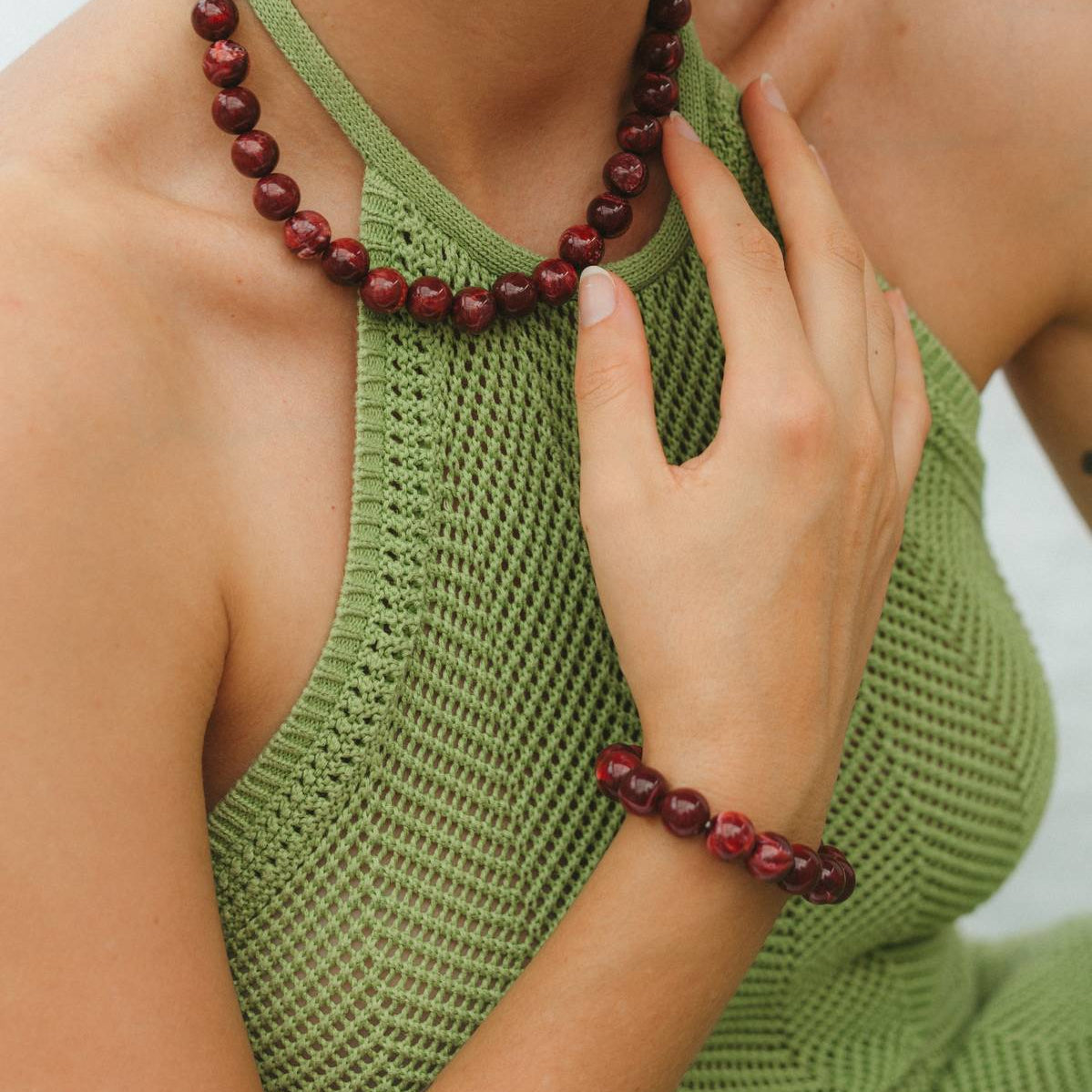 Woman wearing a green sleeveless top and red beaded necklace and bracelet against a neutral background