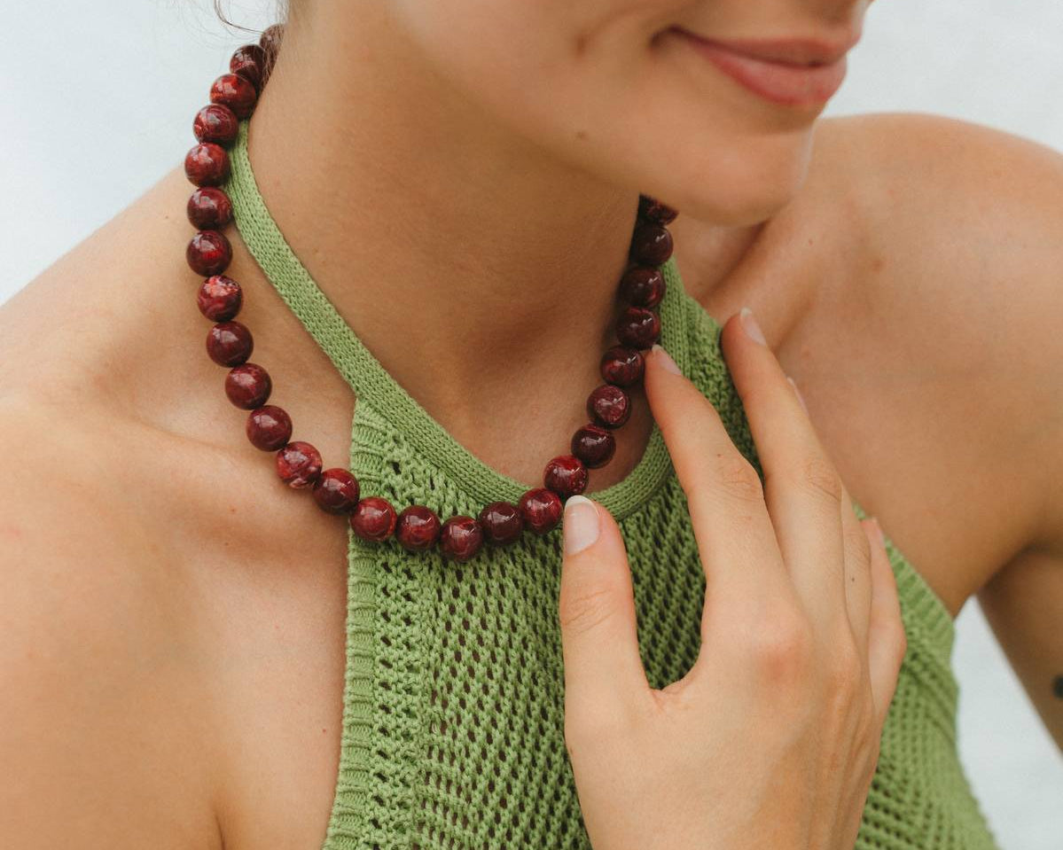 Woman wearing a green sleeveless top and red beaded necklace and bracelet against a neutral background