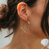 Close-up of an ear wearing a gold hexagonal hoop earring with a neutral background