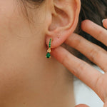 Close-up of an ear wearing a green emerald earring with gold setting, hand touching hair.