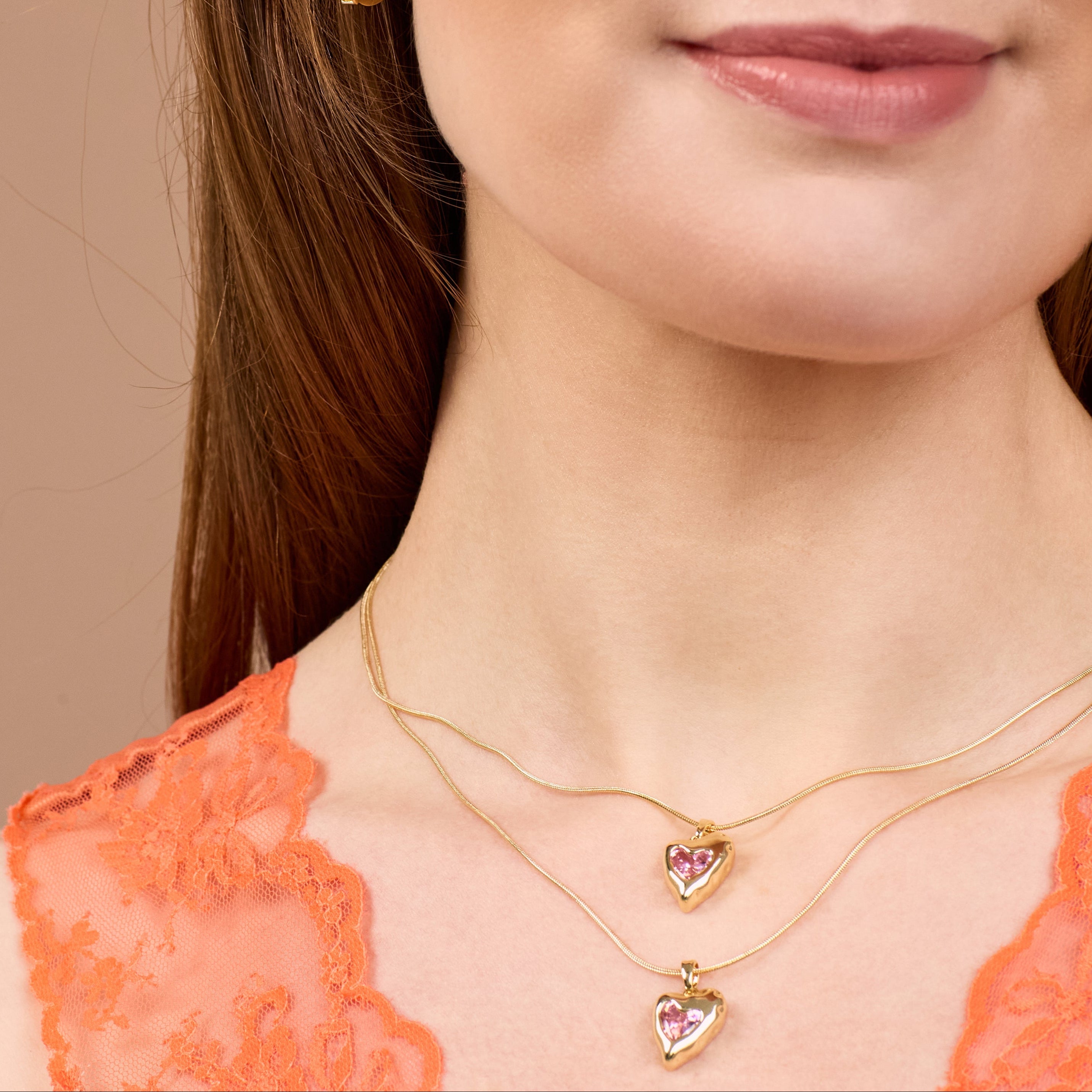 Close-up of a woman wearing two gold necklaces with heart-shaped pendants on a neutral background